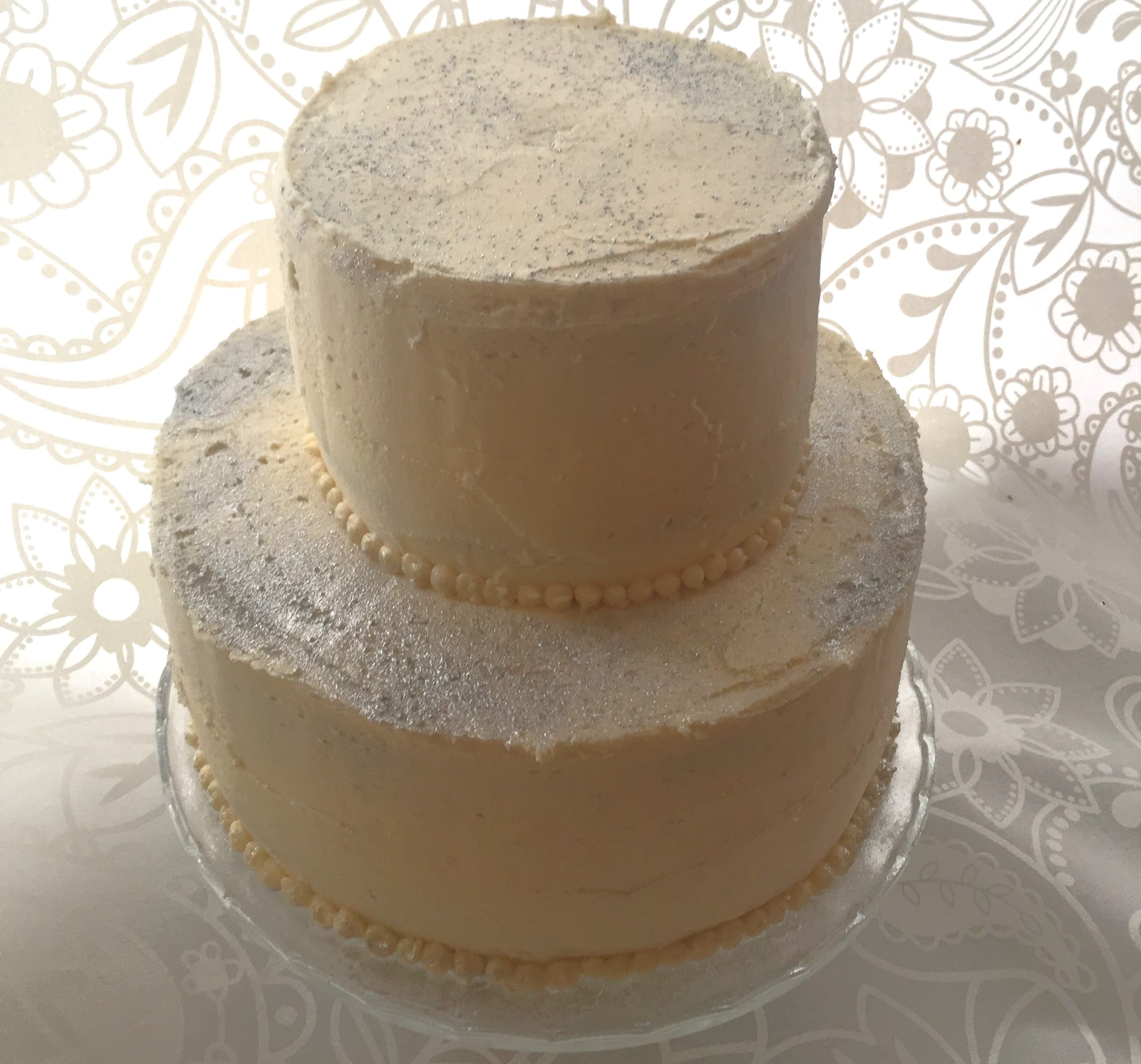 A two-tiered cake with white frosting, lightly dusted with silver sparkles, sits on a glass stand. Each tier is bordered with small pearl-like beads at the base. The background features an intricate floral and paisley pattern in white and beige.